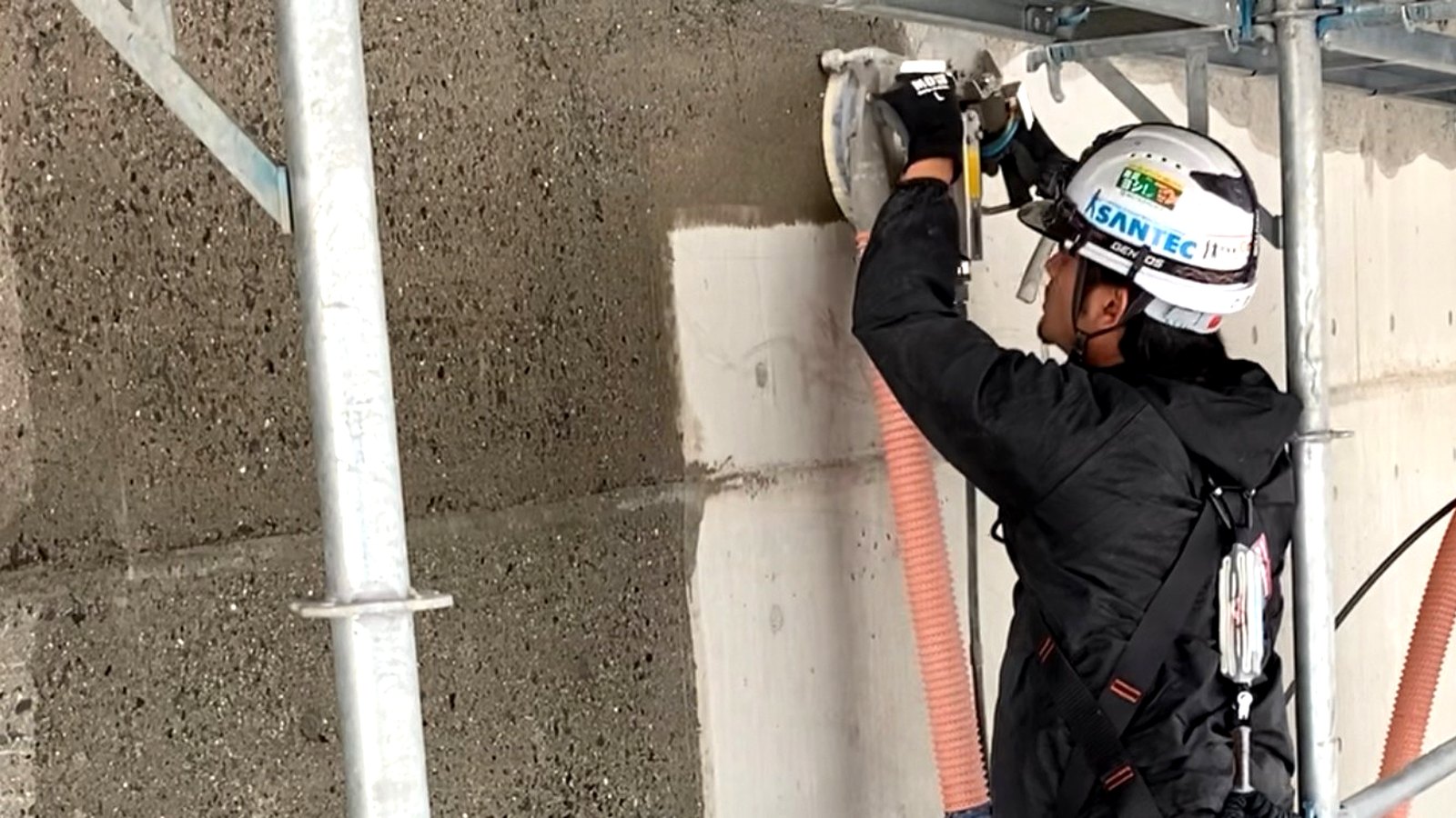 足場の上で作業員が壁面にガンを当てて施工している。SANTECのヘルメットをかぶり、削り終えた部分は周囲より明るい色になっている
