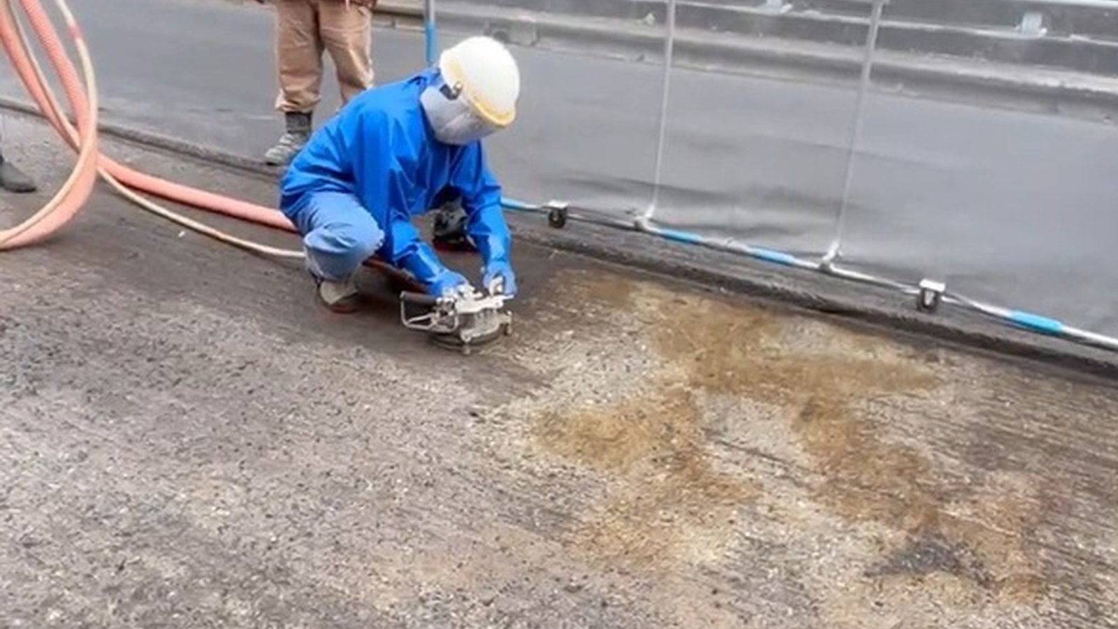 路面にしゃがんだ青い雨具の作業員が、ノズルを組み込んだ台車状の道具を押している。手前の路面は消去済みで色が変わっている